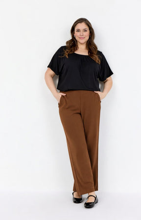 Woman wearing a black top and Wasabi Kufa Trousers in Brown, full length, straight leg trousers with an elasticated waistband with side pockets on a white background