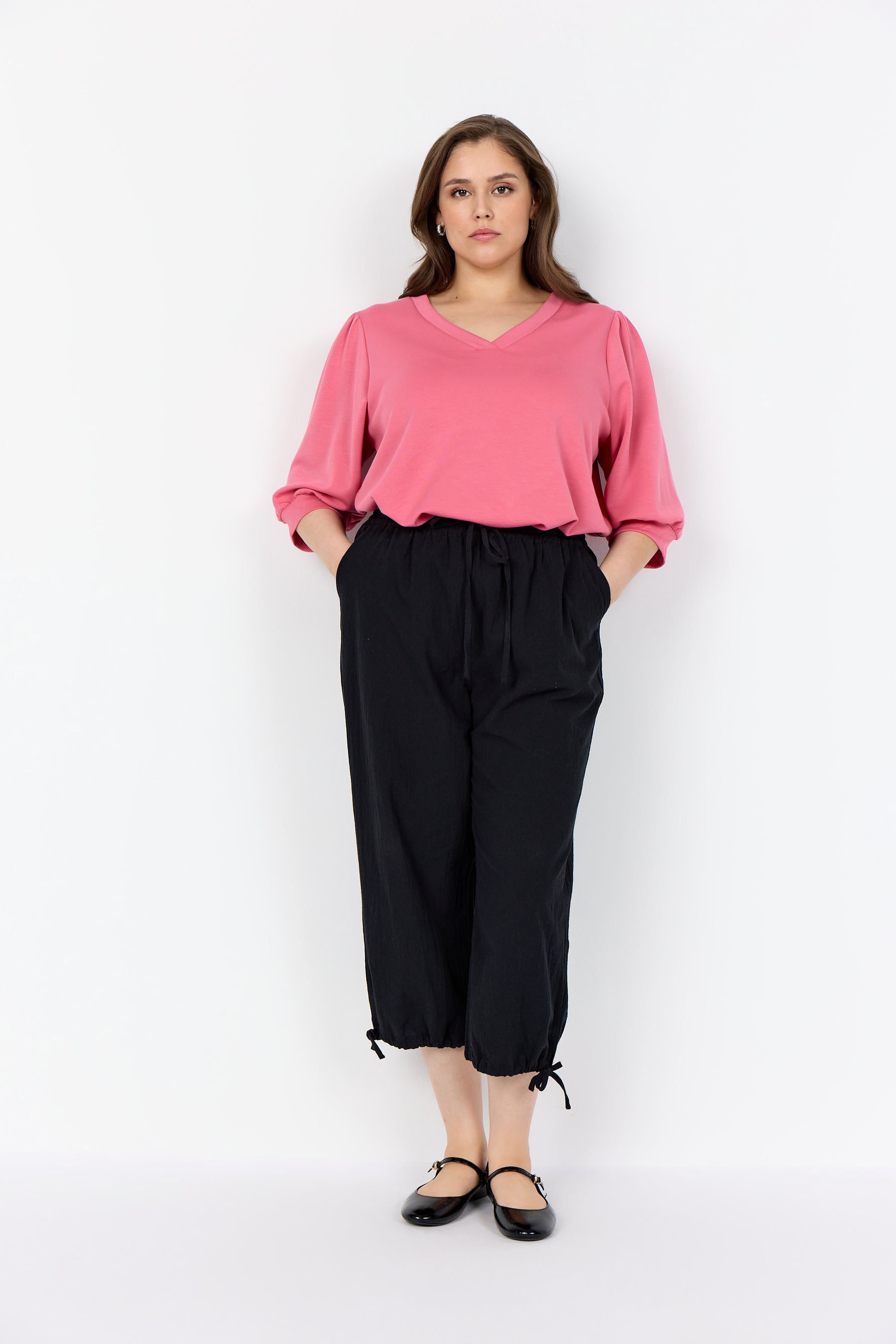 Woman wearing a Wasabi Sabina Blouse in Pink with a V neck, 1/2 length puff sleeves tucked into black pants on a white background