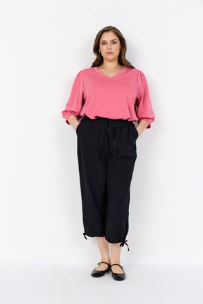 Woman wearing a Wasabi Sabina Blouse in Pink with a V neck, 1/2 length puff sleeves tucked into black pants on a white background