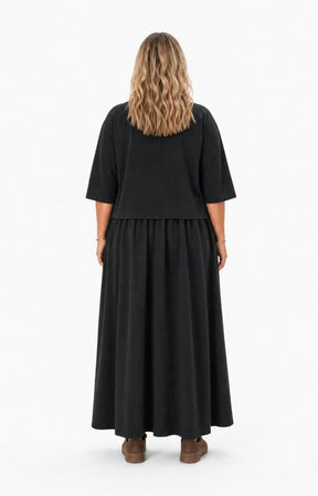 A woman viewed from the back wearing  the Wasabi Sabina Dress in Black, a black two-piece look outfit consisting of a short-sleeve top and a long flared skirt, paired with brown shoes, against a plain white studio background. This image has been created using AI.