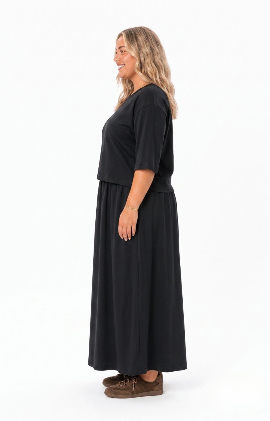 A woman modeling the Wasabi Sabina Dress in Black a black two-piece look outfit consisting of a short-sleeve cropped top and a matching long skirt, paired with brown shoes, against a plain white background. This image has been created using AI.