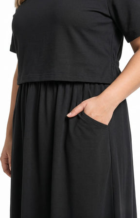 A close-up of a person wearing the Wasabi Sabina Dress in Black, a black dress with a non removable short-sleeve top with one hand in a side pocket against a plain white background. This image was created using AI.