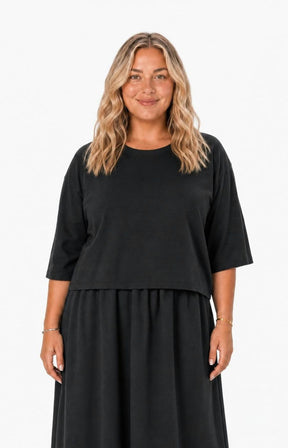 A woman with blonde wavy hair wearing the Wasabi Sabina Dress in Black, a black two-piece look outfit, standing against a plain white background. This image has been created using AI.