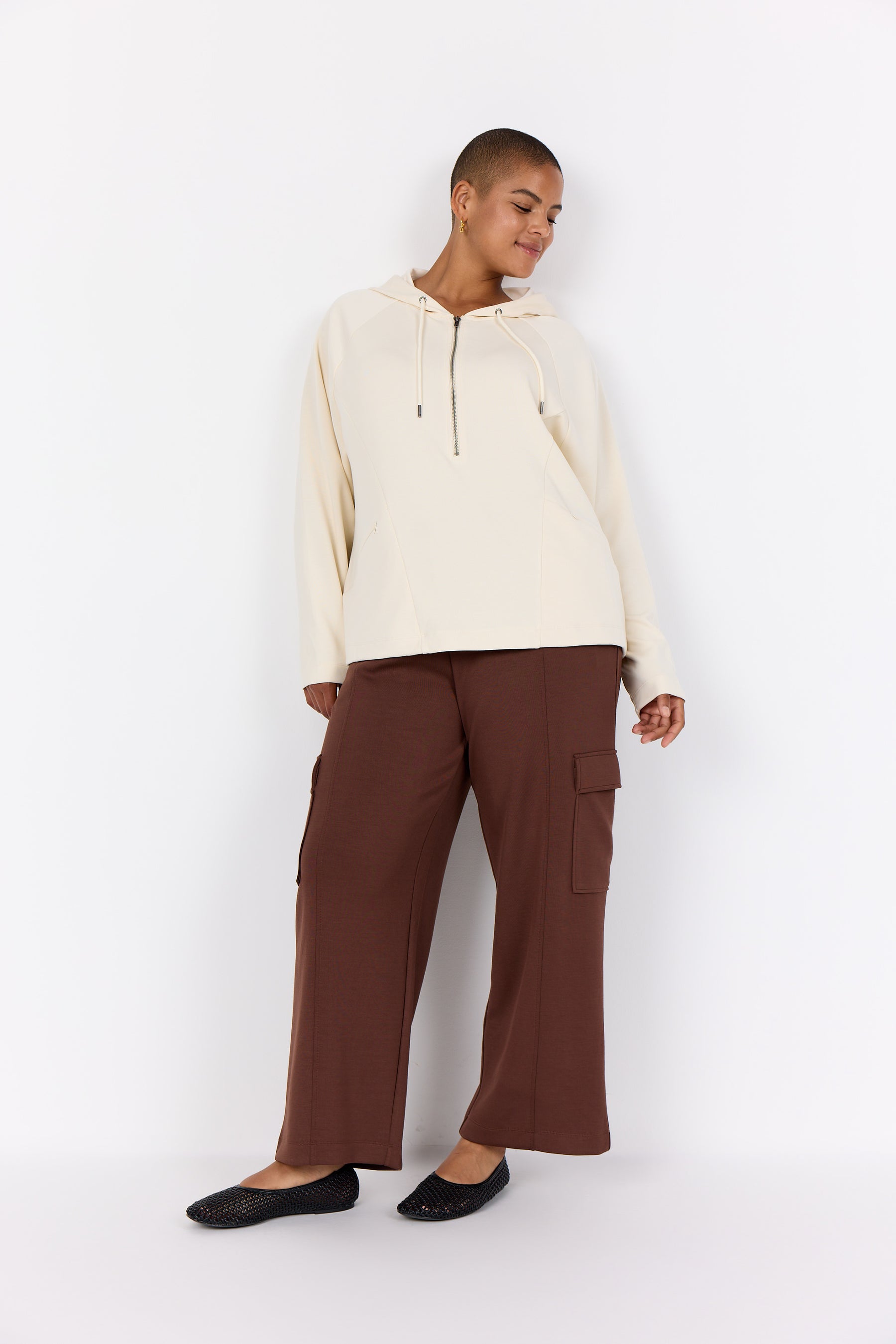 Person wearing a Wasabi Sabina Hoodie in Cream, a hoodie with drawstrings and a 1/2 zip, long sleeves and zipped pockets and brown trousers on a white background