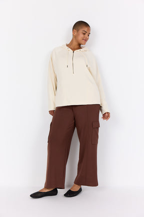 Person wearing a Wasabi Sabina Hoodie in Cream, a hoodie with drawstrings and a 1/2 zip, long sleeves and zipped pockets and brown trousers on a white background