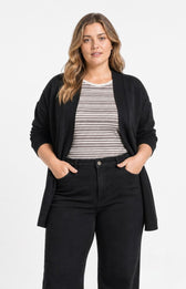 A woman modeling the Wasabi Sabina Pocket Cardigan in Black, a black open-front cardigan over a black and white striped t-shirt and black jeans, standing against a plain white studio background. This image has been created using AI.