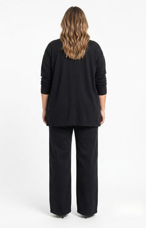 A woman viewed from the back wearing the Wasabi Sabina Pocket Cardigan in Black, a black long-sleeve top and matching black wide-leg pants against a plain white studio background. This image has been created using AI.