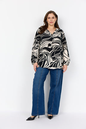 Woman wearing the Wasabi Samena Blouse in Black, a long sleeves, v neck blouse with an abstract black and white design and blue jeans on a white background