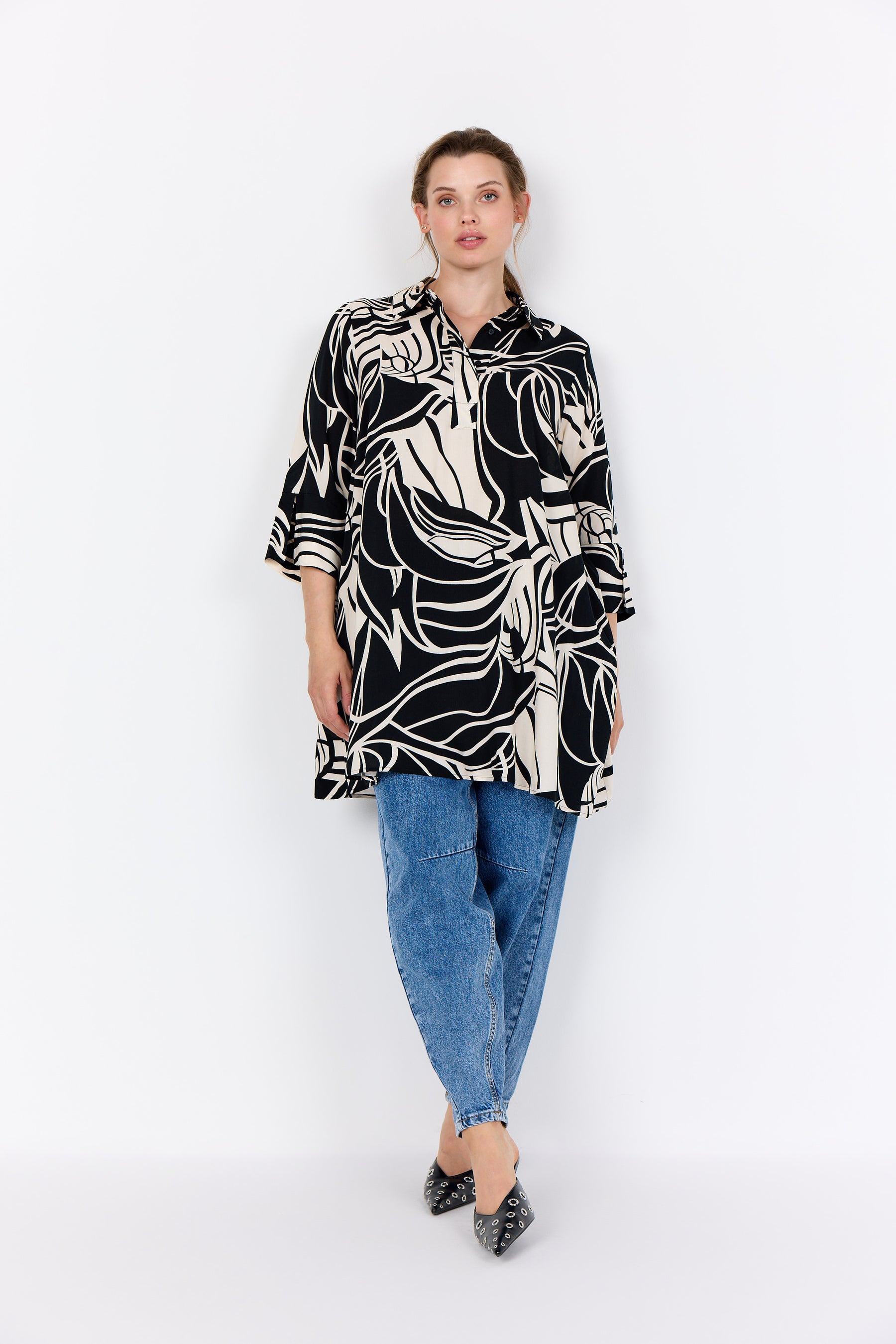 Woman wearing the a Wasabi Samena Tunic in Black, with a shirt collar, button placket, 3/4 length sleeves and an all over print with blue jeans on a white background
