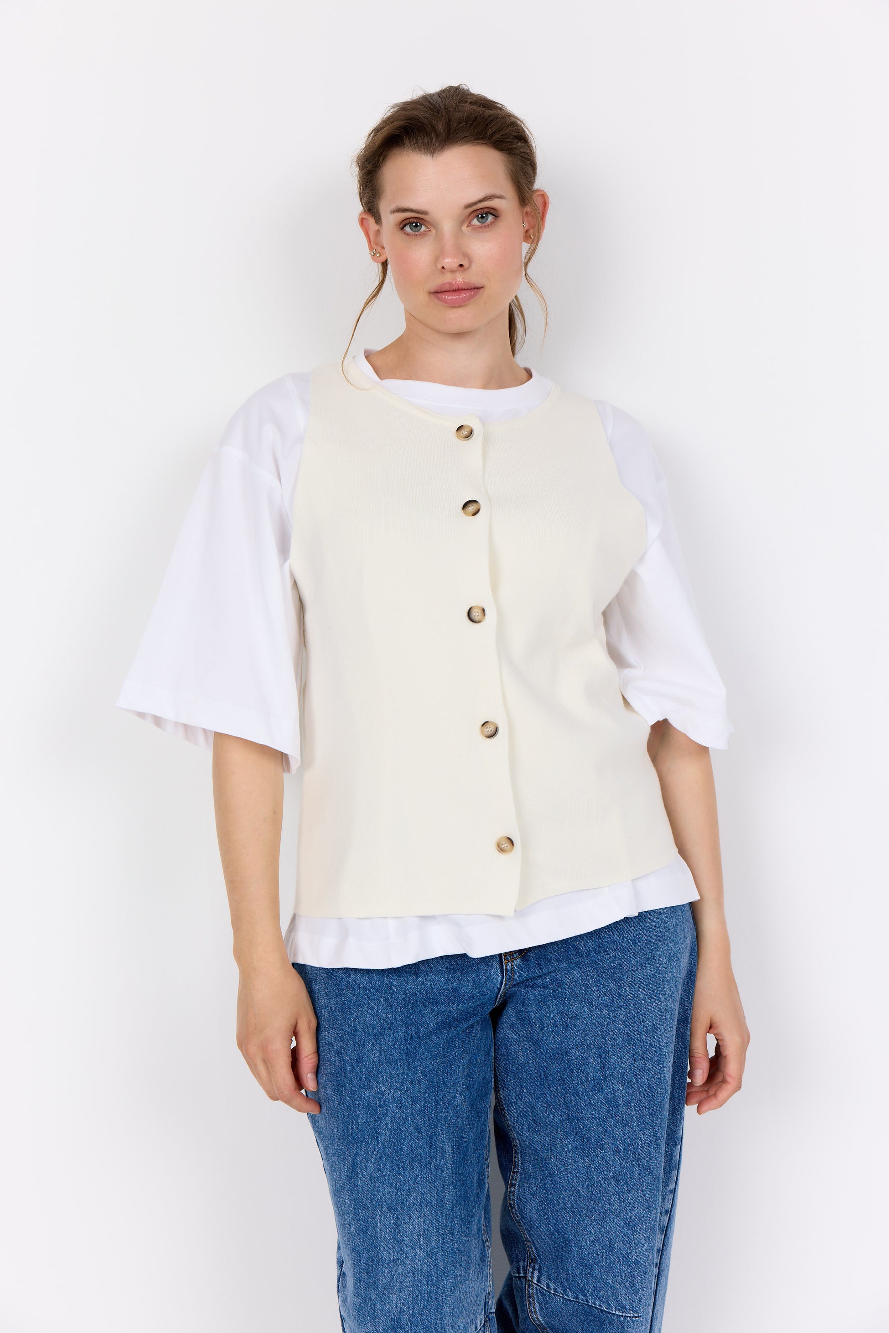 Woman wearing a Wasabi Sikka Waistcoat in Cream with buttons and a round neckline and blue jeans on a white background