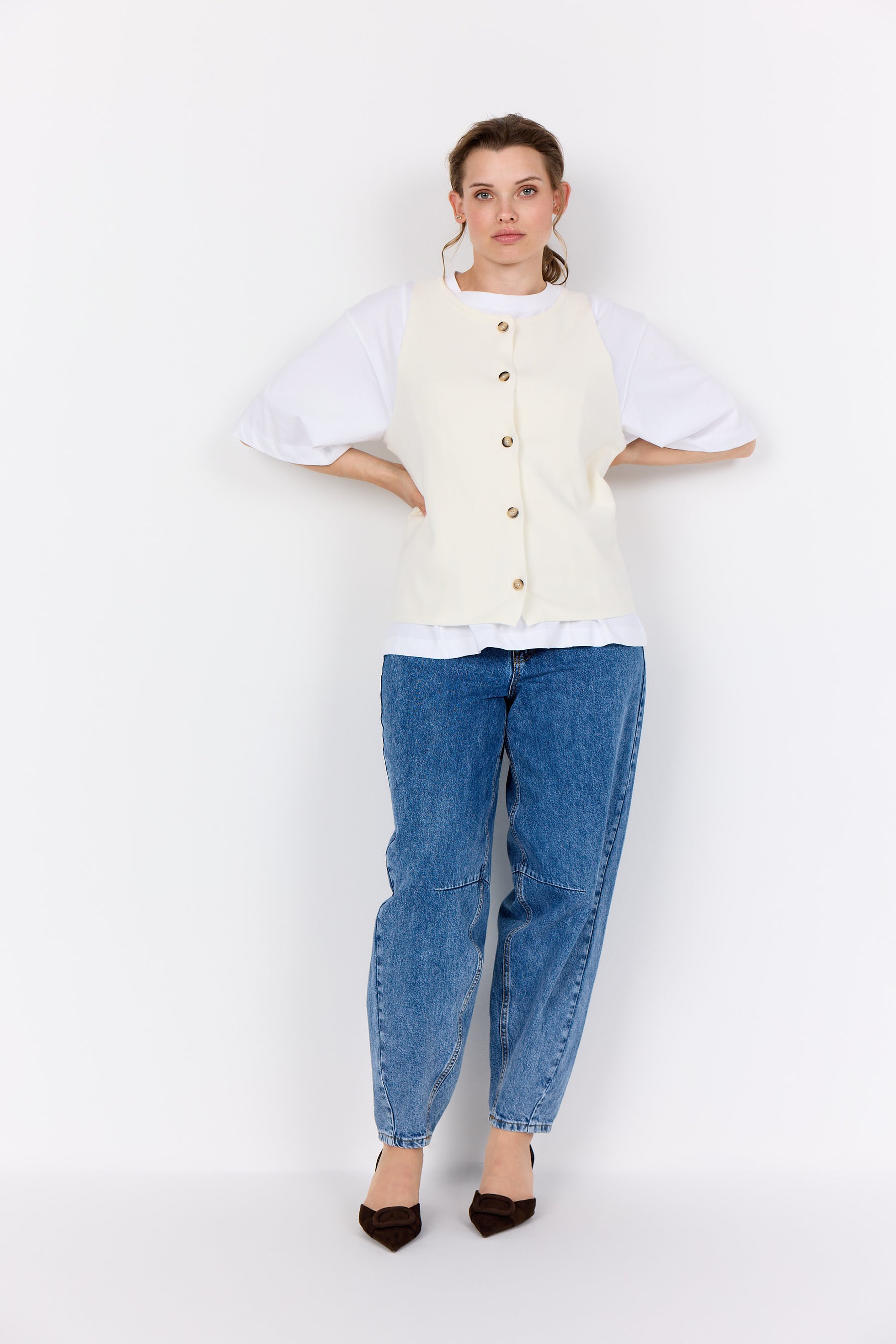 Woman wearing a Wasabi Sikka Waistcoat in Cream, and blue jeans on a white background. The waistcoat has a button up front and round neckline