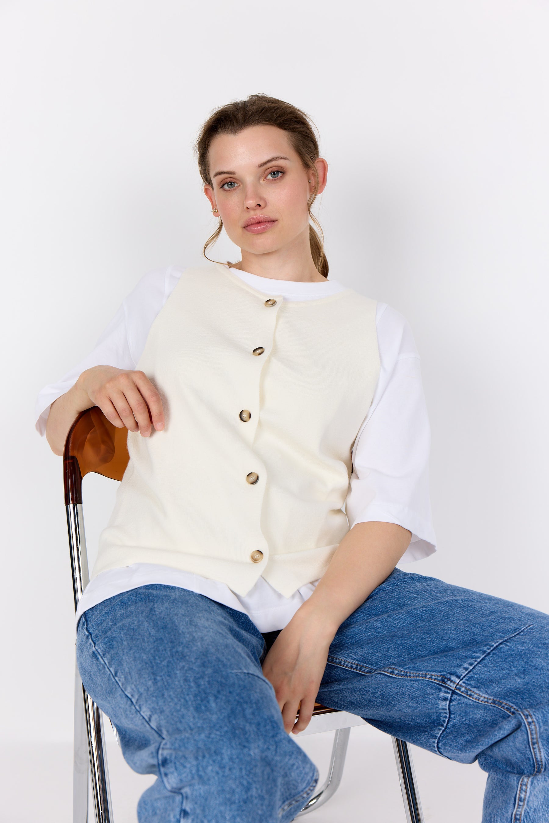 Woman wearing a Wasabi Sikka Waistcoat in Cream over a white shirt and blue jeans, sitting on a chair against a white background.