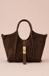 A dark brown suede tote bag with a central gold-tone turn-lock clasp and a matching hanging strap, set against a solid pale pink background.