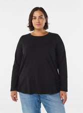 A woman modeling the Zizzi Long Sleeve Top in Black, a black long-sleeve crewneck top with a relaxed fit, paired with blue jeans, against a plain light gray studio background.