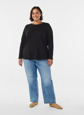 A woman modeling the Zizzi Long Sleeve Top in Black, a black long-sleeve crewneck top and light wash blue jeans with tan flat shoes, standing against a plain white studio background.