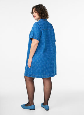 Woman wearing the Zizzi Misha Denim Dress, a blue dress with an all over dot design with an A-line shade and a knee length on a white background