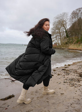 Woman in a black puffer coat standing on a beach with water and trees in the background