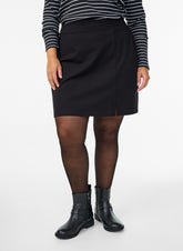 A model wearing the Zizzi Olia Skirt in Black, a black A-line mini skirt, a black and white striped long-sleeve top, sheer black tights, and black leather ankle boots, photographed against a plain white studio background.