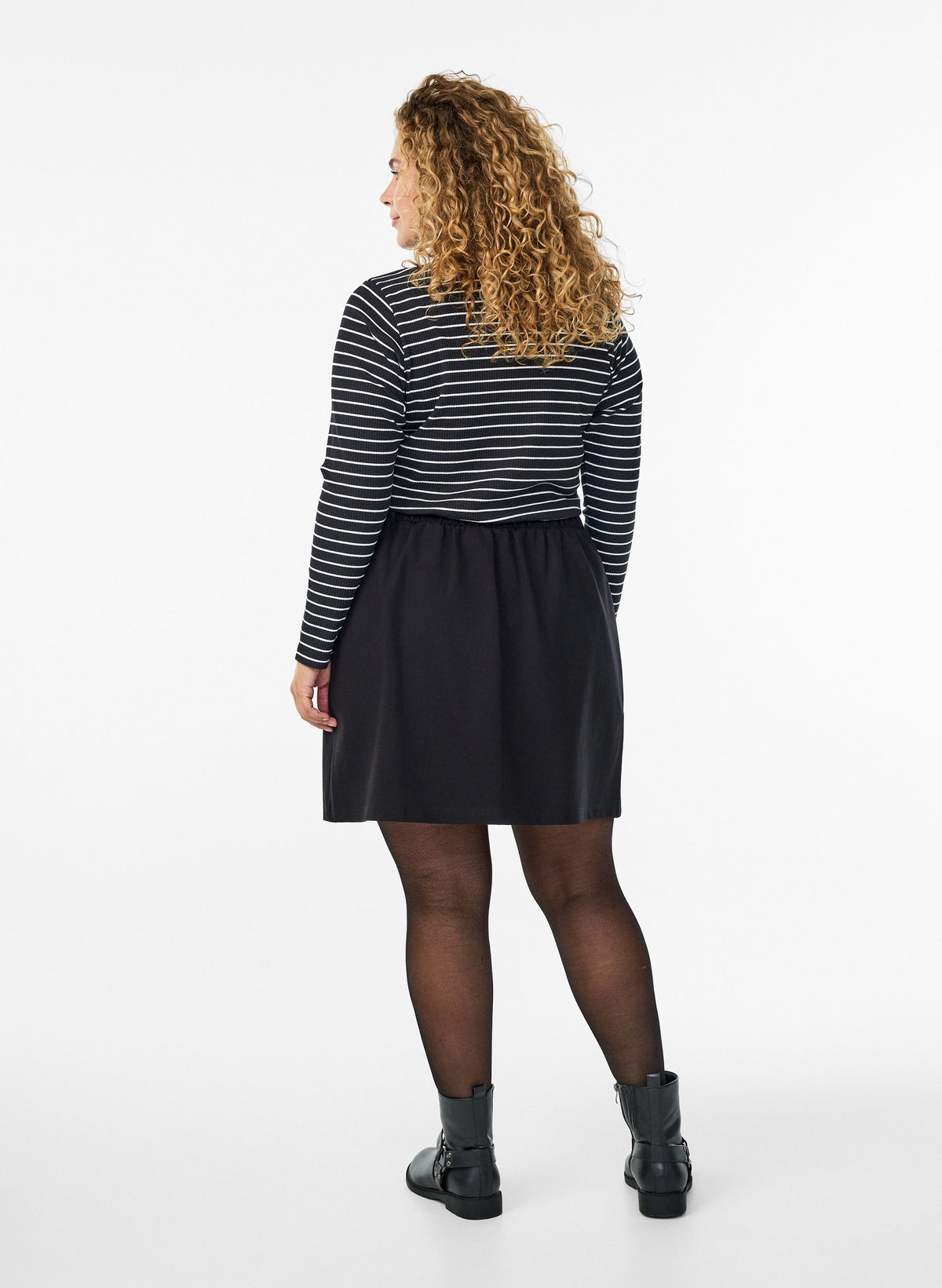 A woman with curly blonde hair is shown from the back wearing a black and white horizontally striped long-sleeve top, a black skirt, sheer black tights, and black ankle boots, against a plain white studio background.