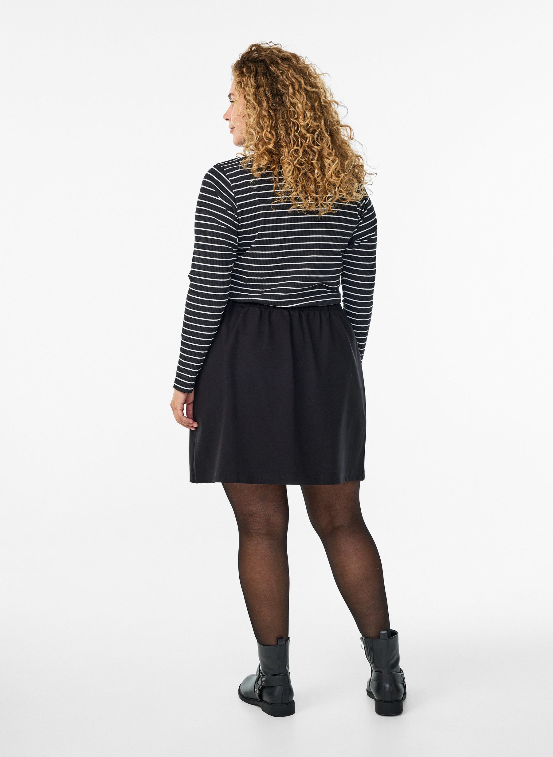 A woman with curly blonde hair is shown from the back wearing a black and white horizontally striped long-sleeve top, a black skirt, sheer black tights, and black ankle boots, against a plain white studio background.