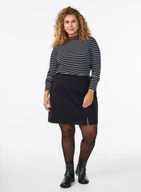 A woman modeling a black and white horizontally striped long-sleeve mock neck top, paired with the Zizzi Olia Skirt in Black, a black skirt, sheer black tights, and black ankle boots, against a plain white studio background.