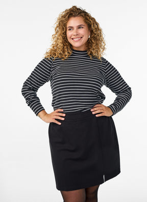 A woman with curly blonde hair models a black and white horizontally striped mock-neck long-sleeve top paired with the Zizzi Olia Skirt in Black, a black skirt, posing against a solid white studio background.