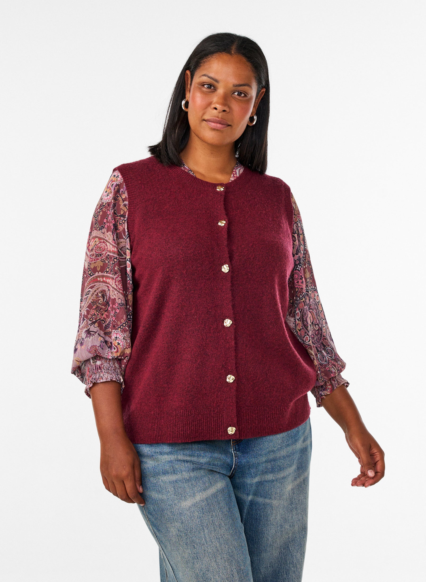 A woman modeling the Zizzi Vista Waistcoat in Windsor Wine, a sleeveless burgundy knit vest with a button-front closure over a patterned long-sleeve blouse and blue jeans, photographed against a plain light gray studio background.