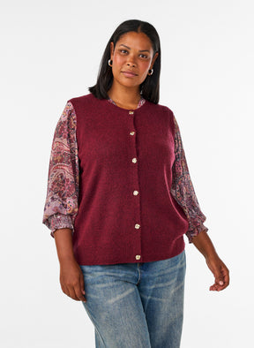 A woman modeling the Zizzi Vista Waistcoat in Windsor Wine, a sleeveless burgundy knit vest with a button-front closure over a patterned long-sleeve blouse and blue jeans, photographed against a plain light gray studio background.