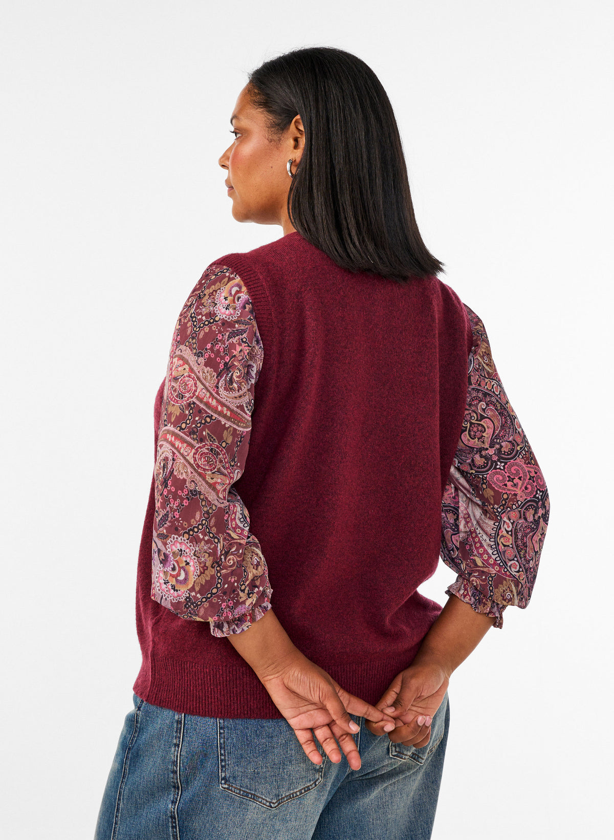 A woman shown from the back wearing the Zizzi Vista Waistcoat in Windsor Wine a burgundy knit sweater vest with patterned puff sleeves, paired with blue jeans, against a plain white studio background. 