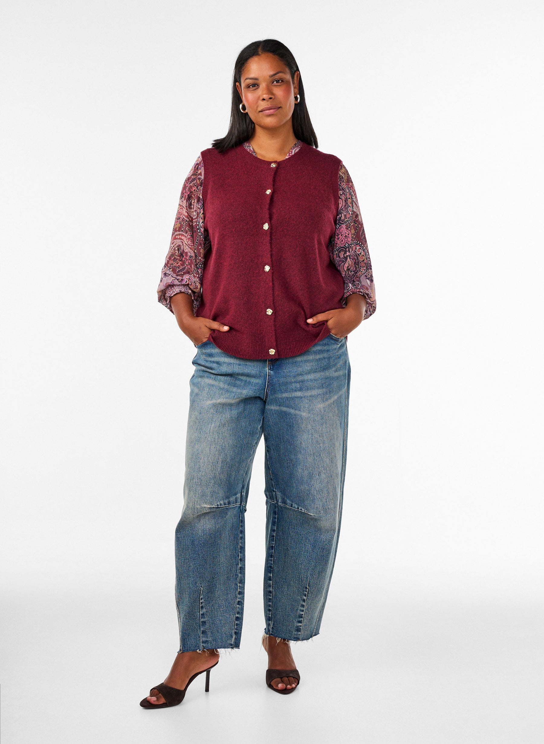 A woman modeling the Zizzi Vista Waistcoat in Windsor Wine a sleeveless burgundy knit vest over a patterned long-sleeve top, paired with wide-leg blue jeans and black heeled sandals, against a plain white studio background.