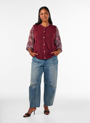 A woman modeling the Zizzi Vista Waistcoat in Windsor Wine a sleeveless burgundy knit vest over a patterned long-sleeve top, paired with wide-leg blue jeans and black heeled sandals, against a plain white studio background.