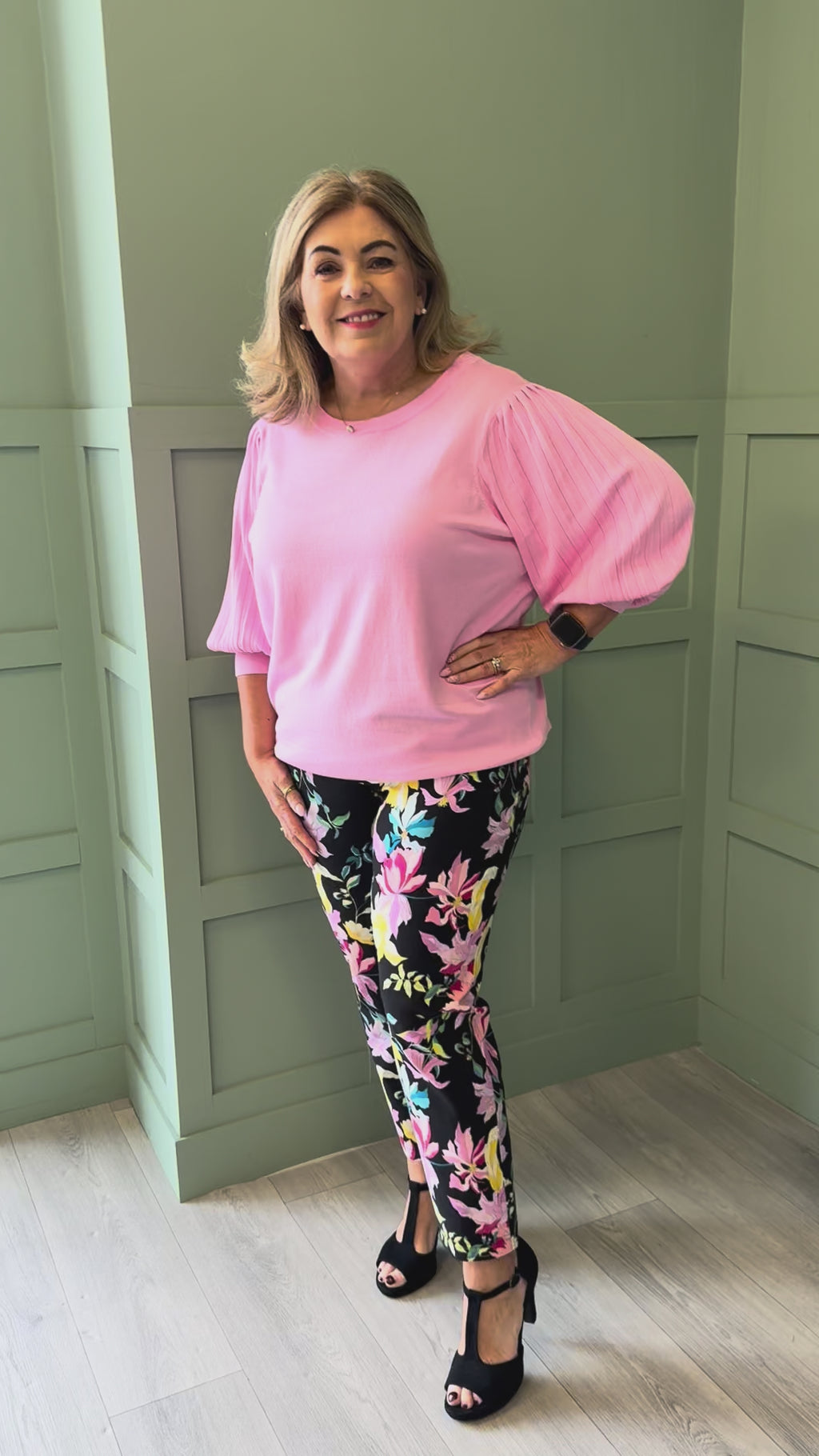 A video of a curvy woman wearing a pink 3/4 sleeve fine knit top and black trousers with a floral print