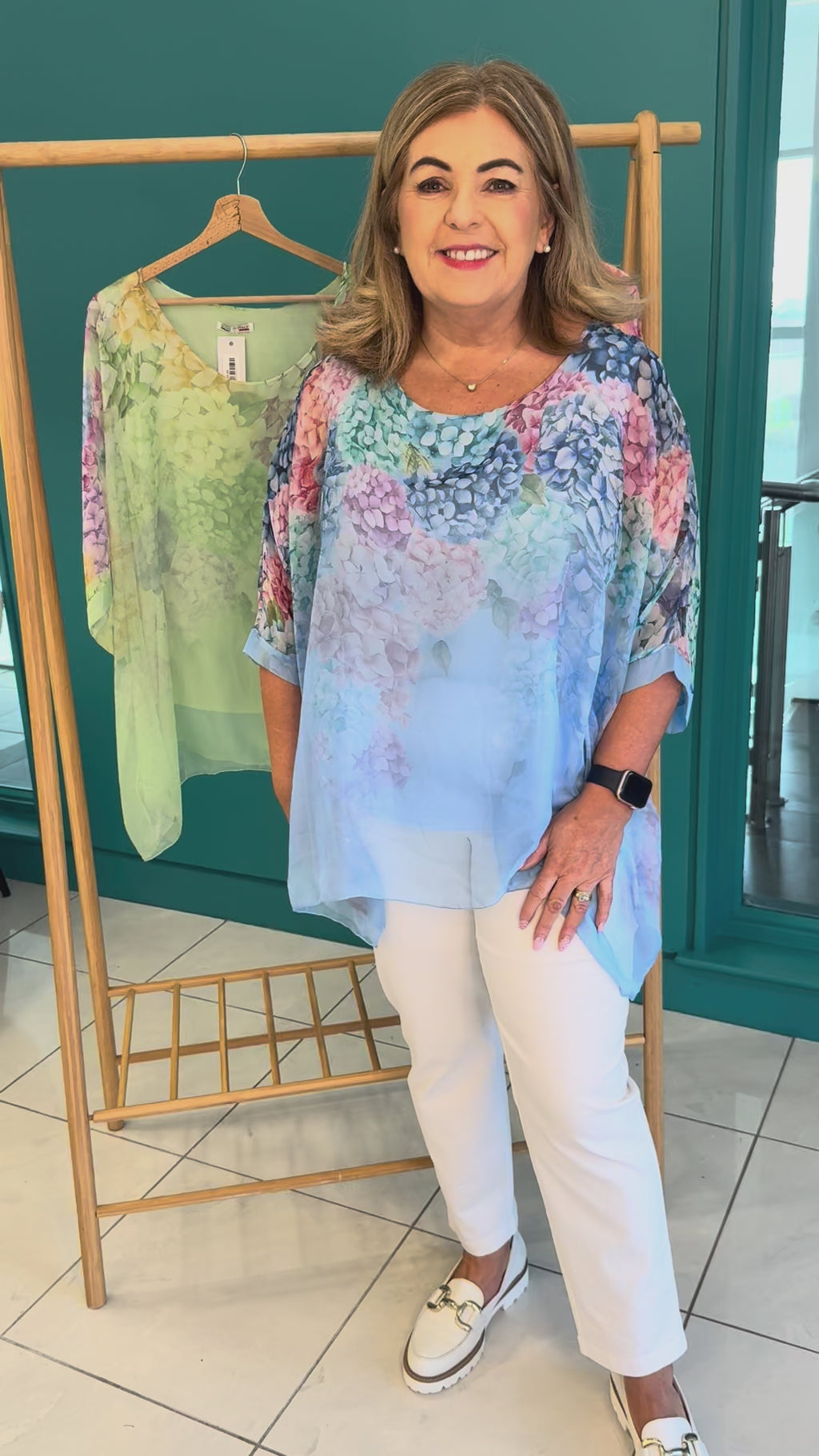 A video of a plus size woman wearing a blue chiffon blouse with a floral print and white trousers. Behind her there's a rail of the same blouses in pink and green
