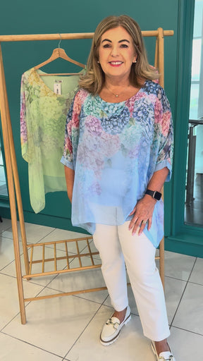 A video of a plus size woman wearing a blue chiffon blouse with a floral print and white trousers. Behind her there's a rail of the same blouses in pink and green