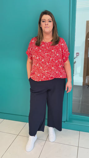 A video of a plus size woman wearing a red blouse with white floral print, navy trousers and white runners.
